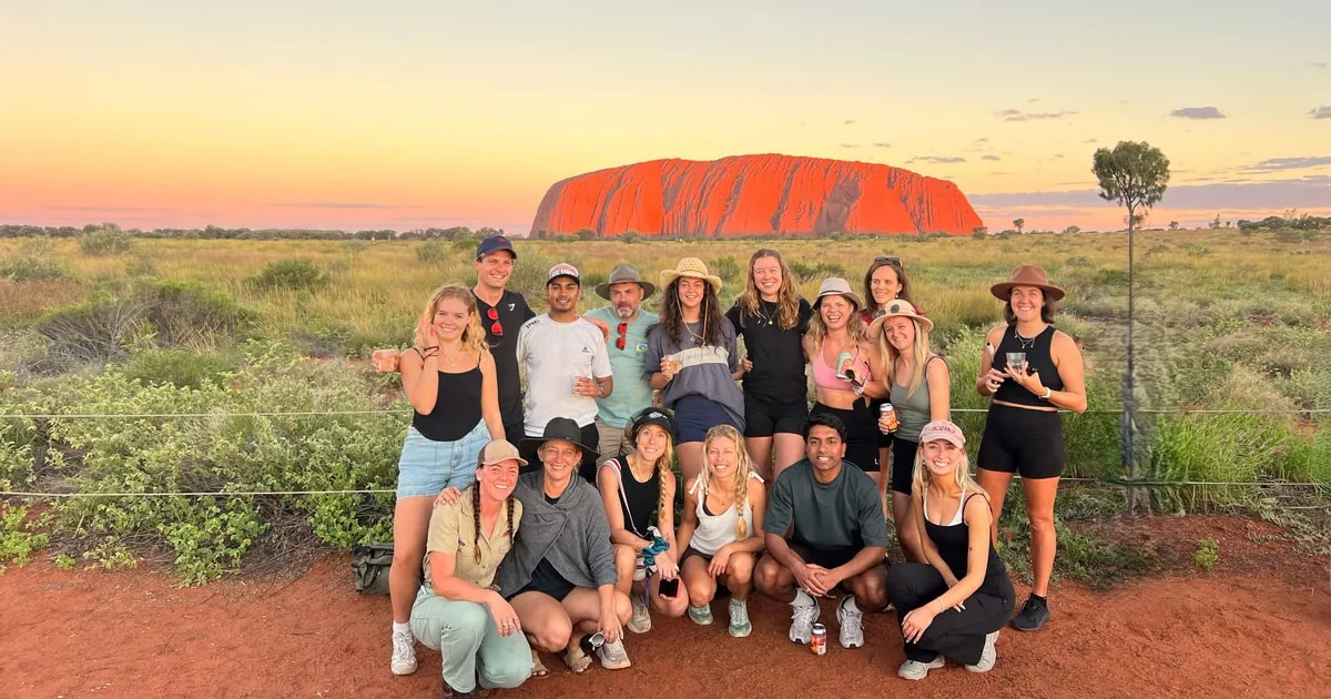 A tour group gathered in the red dirt at sunset with Uluru on the horizon behind them