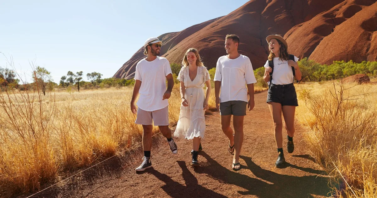 Four young walkers in light clothes on a path through golden grass, with the rock behind them
