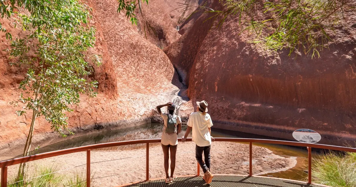 Two walkers at a railing looking at Mutitjulu Waterhole beneath curved red rock walls and gum trees