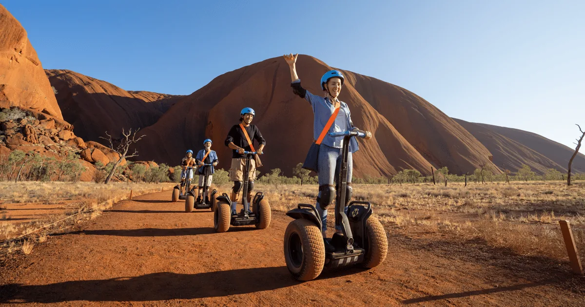 A line of Segway riders on a red track in the morning, with Uluru behind