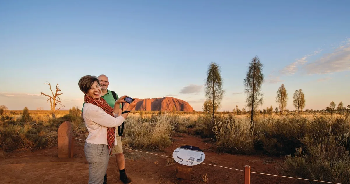 A woman photographing Uluru at sunrise from a fenced viewing area, a man beside her and desert oaks around them