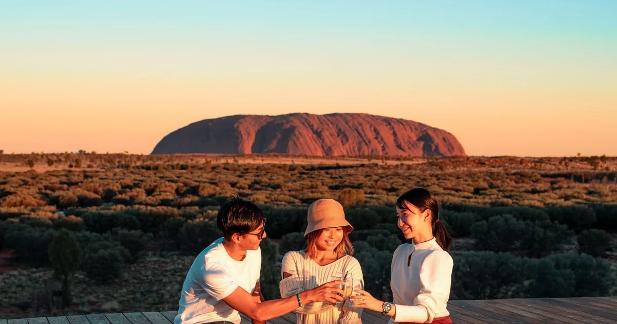 Three people raising glasses on a viewing deck at sunset, with Uluru on the horizon