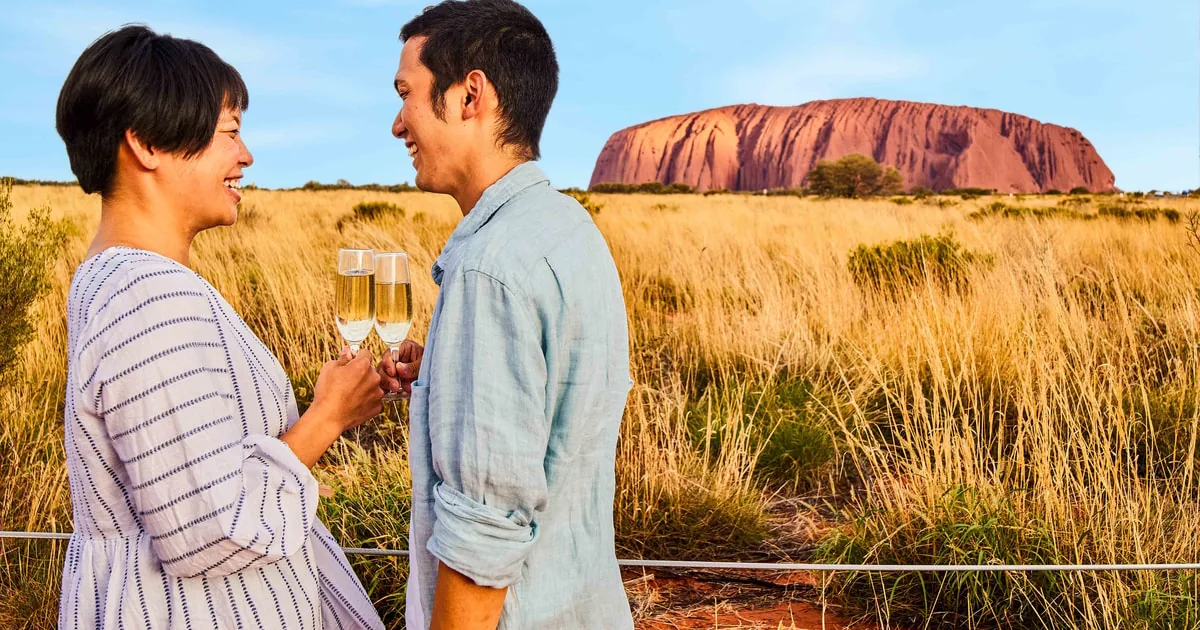 A couple holding glasses of sparkling wine in golden grass, Uluru behind them in the evening light