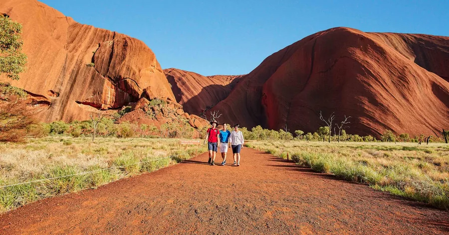 A small group of walkers on a path beneath the steep, fluted red face of Uluru