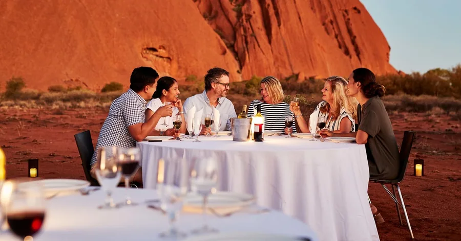 A group dining at a white-clothed table in the desert at dusk, Uluru on the horizon